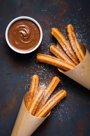 Fried traditional churro sticks with sugar powder cinnamon and bowl of chocolate dip on dark table top viewの写真素材