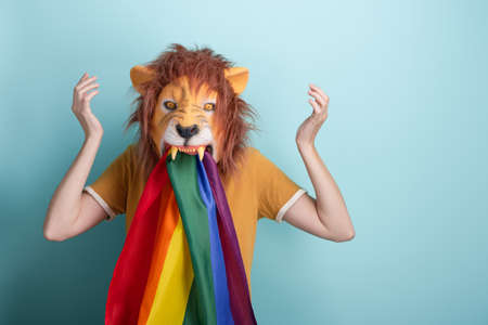 Young woman in lion mask with rainbow pride lgbt flag in mouth disappointed with arms up in air in full disbelief, wtf, annoyed, isolated on blue background with copy-space.の写真素材