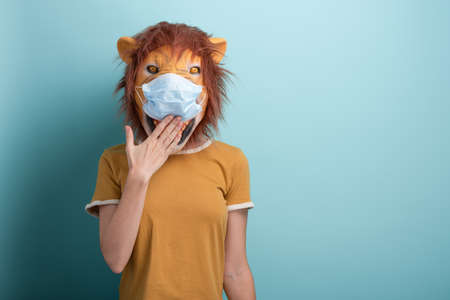 Young woman wearing lion and protection medical mask, covering mouth with hand, isolated on blue background.の写真素材