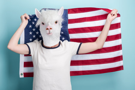 Young woman in alpaca mask hold and wave USA US United States flag in her hands behind head, isolated on blue background.の写真素材