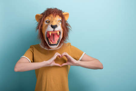 Young woman in lion mask shows love heart affection hand gesture, isolated on blue backgroundの写真素材
