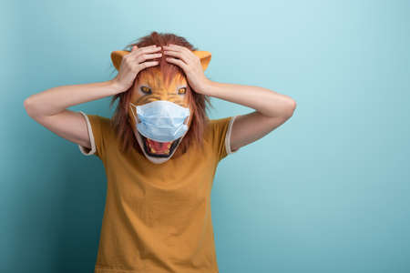 Young woman wearing lion and protection medical mask, shocked, horrified or frustrated with hands on her head, isolated on blue background.の写真素材