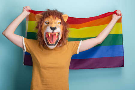 Young woman in lion mask hold and wave pride rainbow lgbt flag in her hands behind head, isolated on blue background.の写真素材