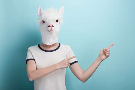 Woman in alpaca mask pointing with index fingers to the blank space, isolated on blue backgroundの写真素材