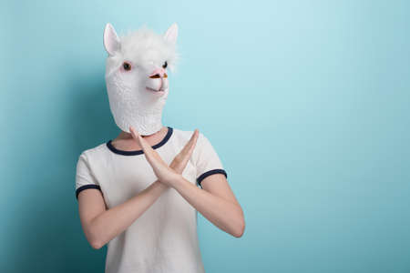 Young woman in alpaca mask hold her hands crossed in stop or cancel sign gesture, isolated on blue background with copy-space.の写真素材