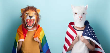 Young woman in lion mask hold rainbow lgbt pride flag wrapped around the shoulders, and other in alpaca mask with USA flag on shoulders, isolated on blue backgroundの写真素材