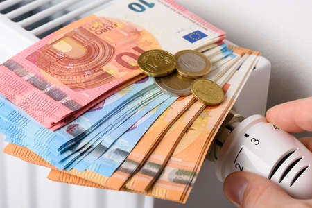 Hand adjusting the valve knob of heating radiator temperature thermostat with stack of euro money banknotes on it. Expensive heating costs and rising energy bill prices for winter cold seasonの写真素材