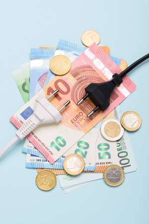 Two electric power plugs on Euro banknotes and coins on blue background. Concept of expensive electricity costs and rise in energy bill prices.の写真素材