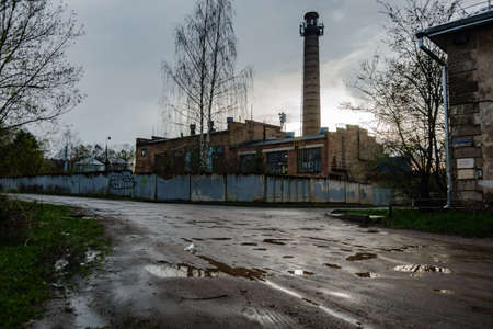 Factory building and brick pipe behind a gray fence, birch, cloudy sky and corner of the houseの写真素材