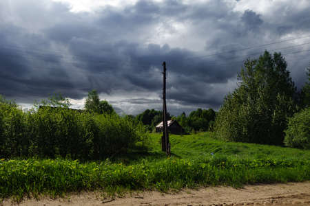 A dirt road and a nearby pole with wires, an abandoned village house, a forest, bushes, a gloomy sky with thundercloudsの写真素材