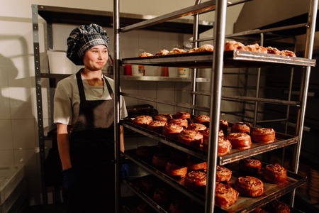 Baker stands near shelves with fresh rolls,baker standing in his bakery in the morning and is baking bread or bunsの写真素材