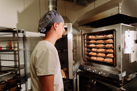 Handsome baker in uniform holding tray full of freshly baked bread at the manufacturing.the Baker...