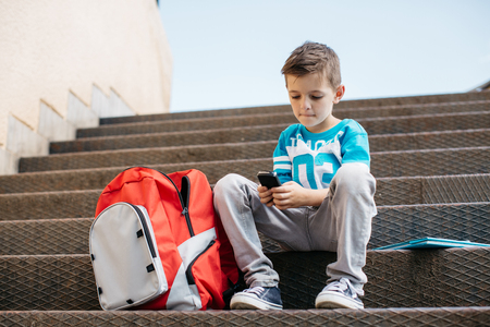 Child sitting outside school and playing games on his cell phoneの写真素材