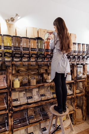 Shopkeeper working in zero waste shop. Shop assistant filling glass jar with red lentils in packaging free grocery store.の写真素材
