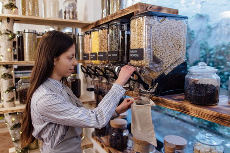 Shop assistant filling reusable bag with dried pasta in organic grocery store. Young shopkeeper working in zero waste shop.の写真素材