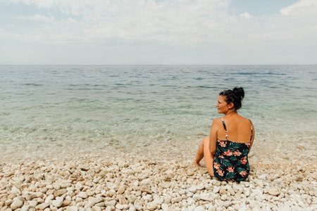 Woman wearing swimwear sitting on stony beach. Summertime relax - back view of woman relaxing at...