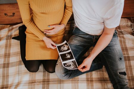 Young couple holding baby scan photos on bed. High angle of young man and woman expecting baby sitting on bed holding baby scans.の写真素材