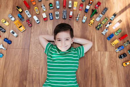 Cute 5 year old boy lying on wooden floor surrounded with car toys. Top view of child wearing green striped t-shirt looking in to camera with car toys around his head.の写真素材