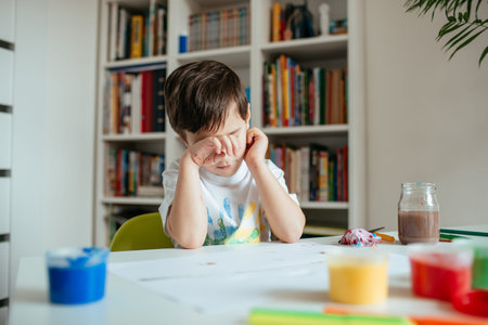 Tired Child Sitting at Table in his Room Rubbing His Eyes. Exhausted Preschooler Boy Loosing Concentration and Getting Tired After Working on his School Painting Assignment.の写真素材
