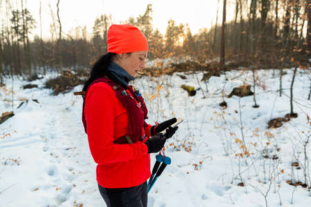 Female Runner Checking Mobile While Running in Woods. Sportswoman With Trekking Poles Holding Cellphone in Hands On Cold Winter Day.の写真素材