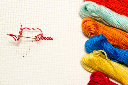 Closeup of cross-stitch: needle with thread on a white canvas with multicolored skeins of thread for...