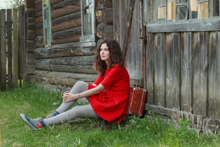 Young woman is sitting on threshold of old log house with vintage suitcaseの写真素材