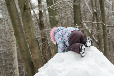 Toddler girl is climbing top of snowy hill in beautiful winter parkの写真素材