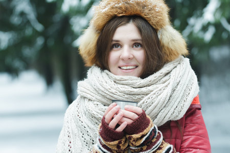 Outdoor winter portrait of cheerful young lady is holding tourist vacuum flask cup with hot drink