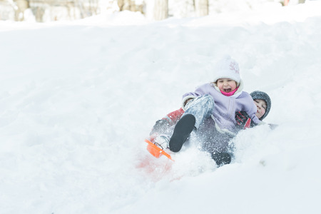 Two children are sliding down snowy hill outdoors on orange plastic modern toboggan for kidsの写真素材