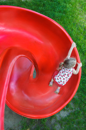 Overhead shot of little blonde girl is sliding down red plastic spiral playground slideの写真素材