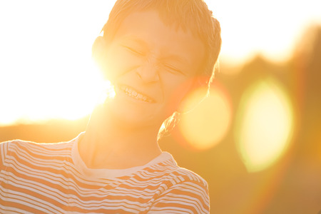 Headshot portrait of screwing up eyes and laughing teenage boy in warm sunset backlit with lens flaresの写真素材
