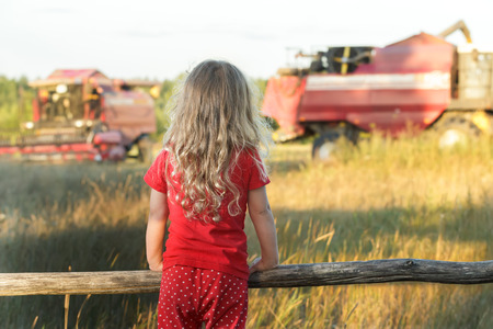 Little farm girl is wearing red polka dot kids pans looking at field with working combine harvestersの写真素材
