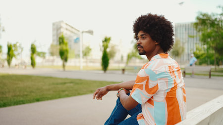 Close-up of young African American man wearing shirt sitting relaxed on park bench looking away....