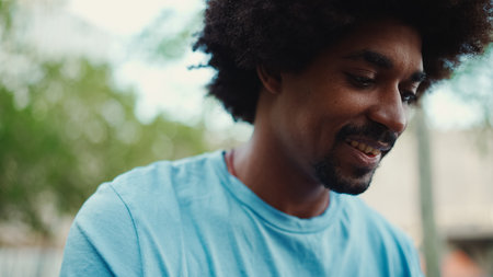 Closeup portrait of young African American man in light blue t-shirt sitting on city park bench and using his smartphone. Man looks at photos, videos in his mobile phone. lifestyle concept.の写真素材