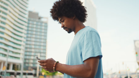 Closeup portrait of young African American man in light blue t-shirt using his smartphone. Man looks at photos, videos in his mobile phone.の写真素材