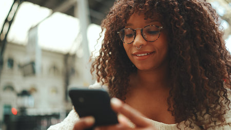 Close-up of a young woman in glasses sits at the station, looks into a smartphone and smiles. Positive woman using mobile phone outdoors in urban background.の写真素材