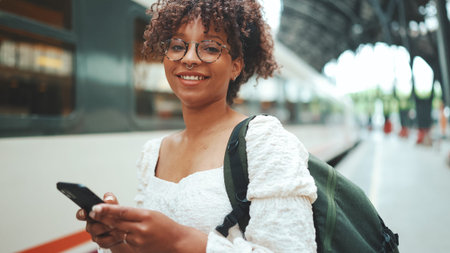Close-up portrait of a young woman in glasses with a smartphone and backpack stands at the station...