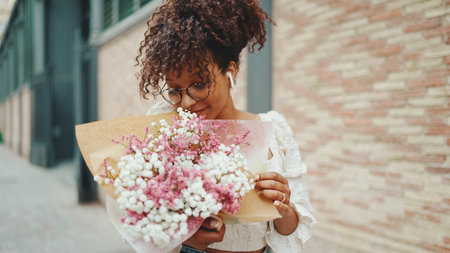 young woman in glasses walks with a bouquet of flowers along the street of the old city and listens to music in wireless headphonesの写真素材
