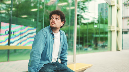 Young bearded man in denim shirt sits at bus stop studying city transport timetables and looking...