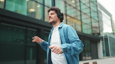 Emotional young bearded man in denim shirt walks down the street in headphones listening to music...
