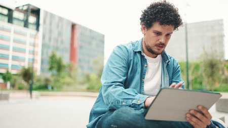 young bearded man in denim shirt sitting and using tablet outdoors on modern buildings backgroundの写真素材