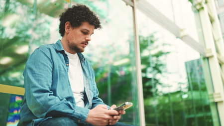 Close-up, young bearded man in denim shirt sits at bus stop waiting for bus and uses mobile phone on...