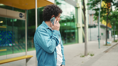young bearded man in denim shirt stands at bus stop waiting for bus and talking on mobile phone on...