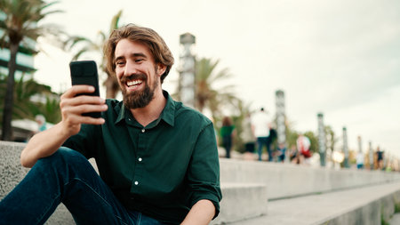 Close-up portrait of a man with a beard recording a voice message on the embankment background. Close up of a young hipster male using a mobile phoneの写真素材