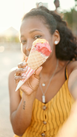 Closeup of smiling young woman with long curly hair with ice cream in her hands making a video call...