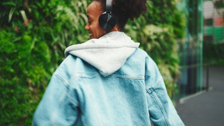 Closeup, smiling African girl wearing denim jacket, in crop top with national pattern walks in city park listening to music on headphones and dancing on green trees background.の写真素材