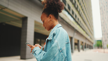 Close-up African girl wearing denim jacket, in crop top with national pattern goes on the street of modern city and holding smartphone looking at phone screen look for address with electronic map in cityの写真素材