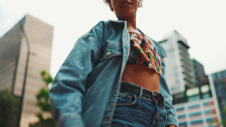 Closeup, smiling African girl with ponytail wearing denim jacket, in crop top with national pattern listening to music on headphones and dancing outdoors.の写真素材