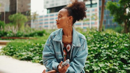 African girl with ponytail wearing denim jacket leaving voice message on mobile phone against modern...