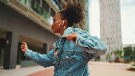 Closeup, smiling African girl with ponytail wearing denim jacket, in crop top with national pattern listening to music on headphones and dancing outdoors.の写真素材